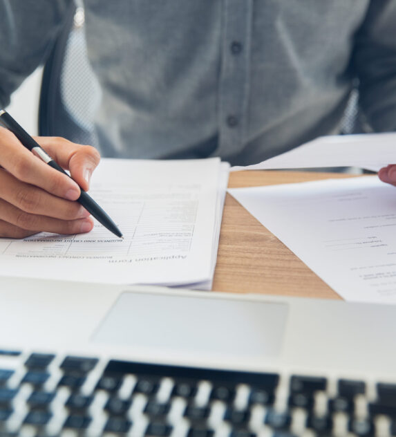 Unrecognizable businessman sitting at table holding ballpoint pen and papers and checking documents in office